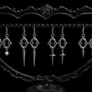 Peut inclure: Quatre paires de boucles d'oreilles argentées suspendues sur un présentoir noir. Chaque paire présente un haut en forme d'anneau ovale orné identique. Les pendentifs varient, incluant des perles blanches, des pointes et des étoiles.