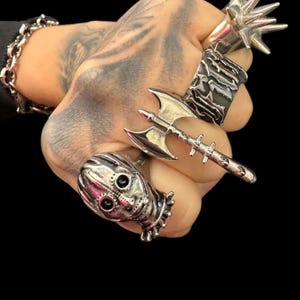 Può includere: Un set di quattro anelli d'argento con un teschio, un'ascia, un anello con punte e una fascia testurizzata. Gli anelli sono indossati su una mano con tatuaggi.