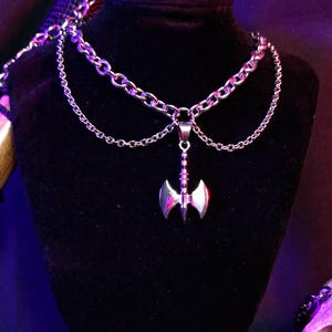 May include: A silver chain necklace with a double chain design and a silver axe pendant. The necklace is displayed on a black velvet stand.