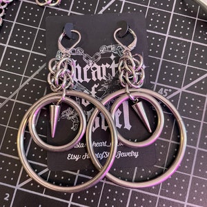 May include: A pair of silver hoop earrings with a smaller hoop inside. Each earring has a small spike in the center of the smaller hoop. The earrings are displayed on a black card with the text "Heart of Steel Jewelry" printed on it.