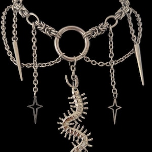 May include: Silver-toned necklace with a central ring and multiple chains. The design includes a centipede pendant, star-shaped charms, and spike accents. The necklace is set against a black background.