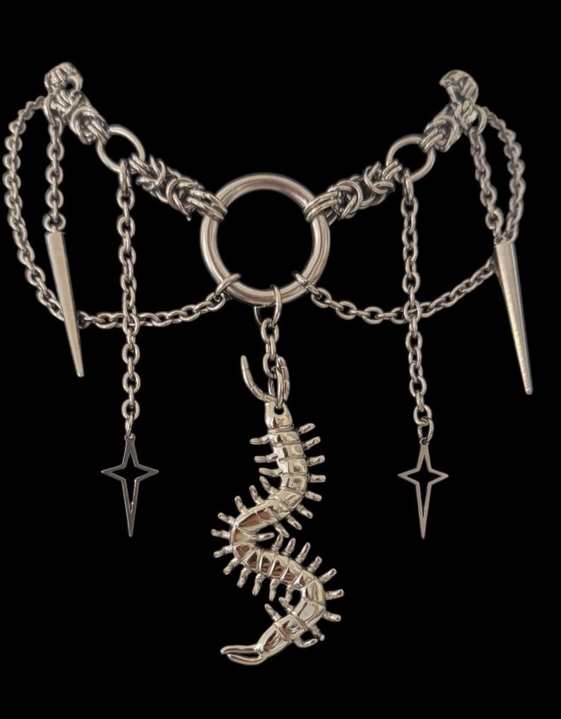 May include: Silver-toned necklace with a central ring and multiple chains. The design includes a centipede pendant, star-shaped charms, and spike accents. The necklace is set against a black background.