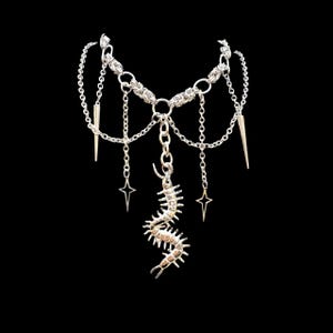 May include: Silver-toned necklace with a layered design. The necklace features a chain base with dangling chains, star charms, and spike pendants. A centipede-like pendant hangs from the center. The jewelry is set against a black background.