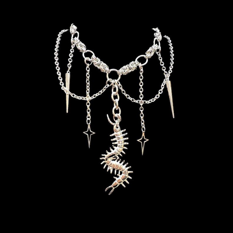 May include: Silver-toned necklace with a layered design. The necklace features a chain base with dangling chains, star charms, and spike pendants. A centipede-like pendant hangs from the center. The jewelry is set against a black background.