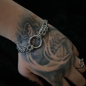 May include: A silver chain bracelet with a large, round, silver ring connecting the two ends. The bracelet is on a person's wrist.
