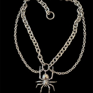 May include: A silver chain necklace with a silver spider pendant. The necklace has two chains, one thicker than the other.