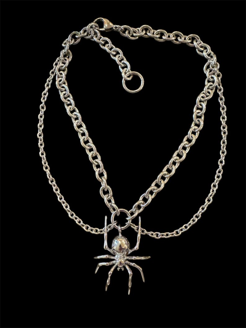 May include: A silver chain necklace with a silver spider pendant. The necklace has two chains, one thicker than the other.