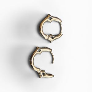 May include: A pair of small, circular hoop earrings crafted from a polished metallic material. Each earring features a distinct, pointed decorative element at the top and bottom, creating a symmetrical, angular profile with a hinged closure mechanism.