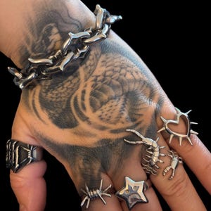 May include: A silver-toned metal chain bracelet and a collection of six silver-toned metal rings. The rings feature various designs including a barbed wire motif, a star with a sad face, a centipede, a crab, and a heart with spikes.