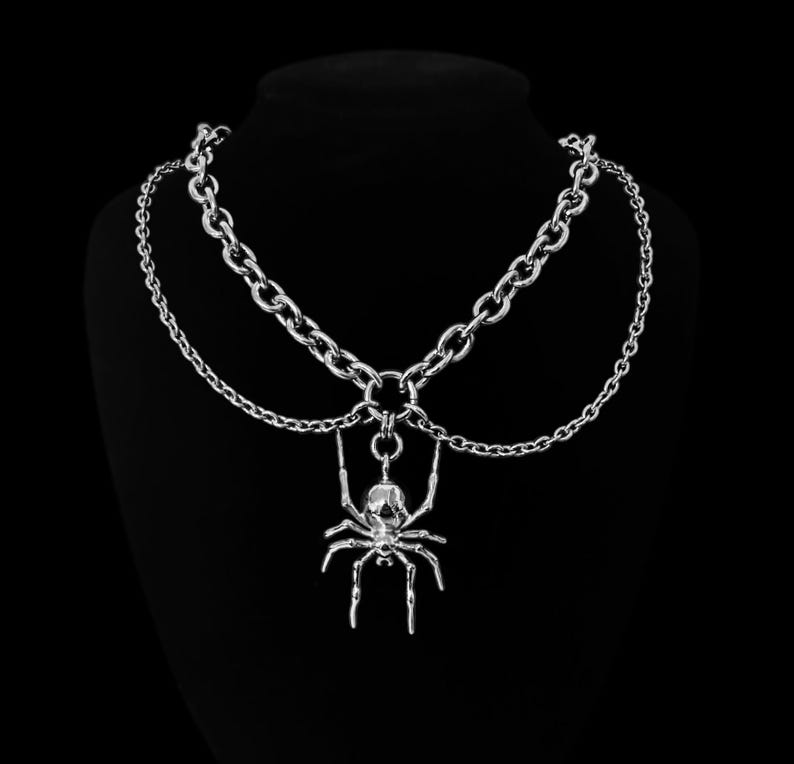 May include: A silver chain necklace with a dangling spider pendant. The necklace is layered with two chains, one thicker than the other.