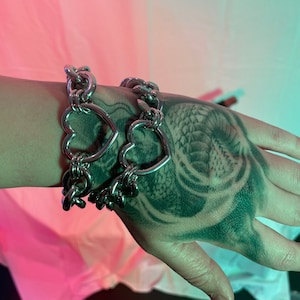 May include: A silver chain bracelet with heart-shaped links. The bracelet is worn on a hand with a tattoo.