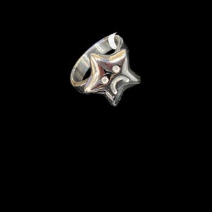 May include: A polished silver-toned ring featuring a five-pointed star design. The star face displays two small circular indentations for eyes and a curved, downward-facing line for a mouth, creating a sad facial expression on the metal surface.