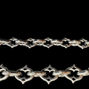 May include: Two silver chains with decorative, gothic-style links against a black backdrop. The links feature a repeating pattern of pointed arches and diamond shapes, creating an ornate design. These chains are likely jewellery.
