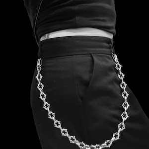May include: A silver-toned metal wallet chain featuring a repeating ornate cross-like link design. The chain is attached to the waistband of black trousers, draping in a curved shape along the side of the garment, creating a decorative accessory.