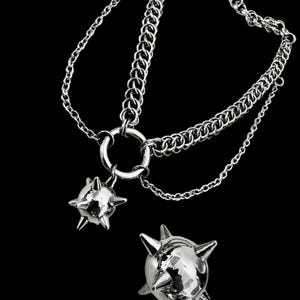 May include: A silver chain necklace with two spiked ball pendants. The chain is made of two different styles of links, one a simple chain and the other a more intricate design. The spiked balls are both round and have a shiny finish.