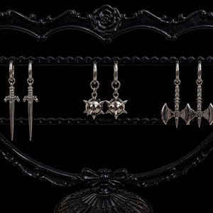 May include: Three pairs of silver-toned metal hoop earrings hanging on a black heart-shaped display stand. The earrings feature dangling charms shaped like swords, spiked maces, and double-headed battle axes with intricate metallic detailing.