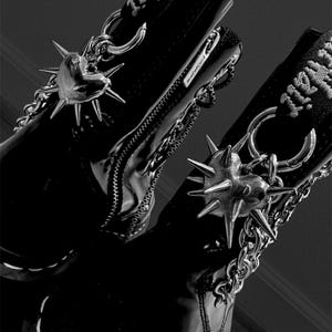 Puede incluir: Un par de botas negras de charol con detalles de cadena plateada y pinchos en forma de corazón. Las botas tienen una cremallera lateral y una correa negra con texto blanco que dice "All Hell."