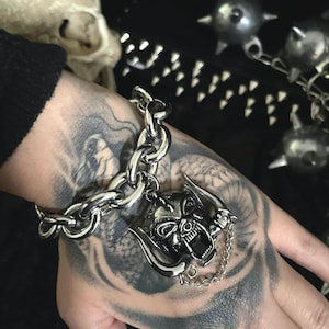May include: A silver chain bracelet with a skull pendant featuring horns and a chain detail. The bracelet is on a hand with a tattoo.