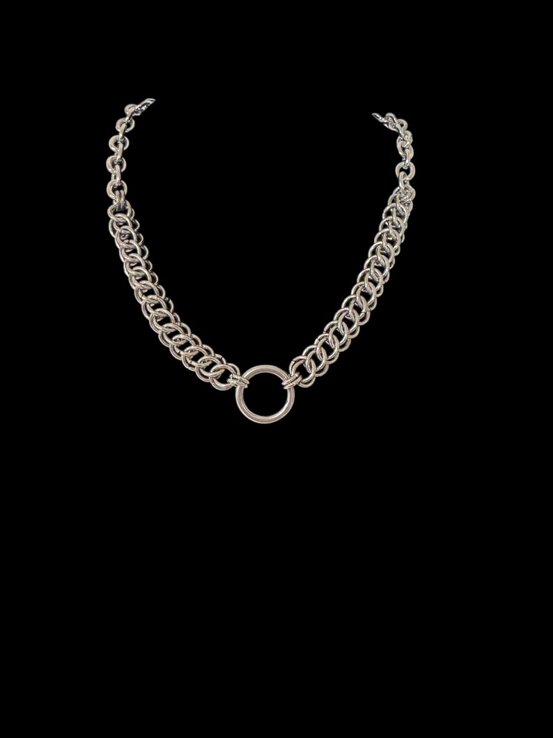 May include: Silver-toned chain necklace with a large circular pendant. The necklace features a double-strand chain design with a series of interlocking links. The pendant is a simple, polished ring. The jewelry is set against a black background.