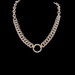 May include: A silver-toned necklace with a large circular pendant. The necklace features a double-strand chain design, with a smaller chain connecting to the clasp. The background is black.