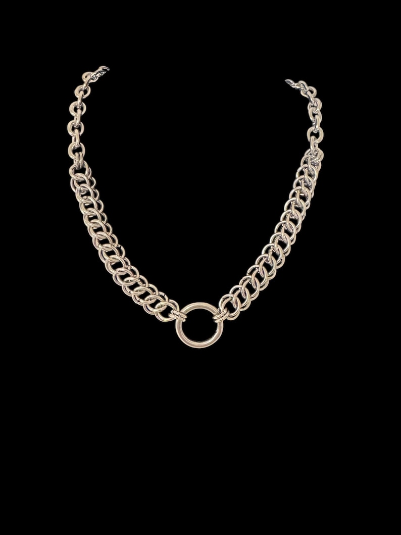 May include: A silver-toned necklace with a large circular pendant. The necklace features a double-strand chain design, with a smaller chain connecting to the clasp. The background is black.