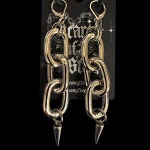 May include: Silver chain link earrings with a pointed spike at the bottom. The earrings are displayed on a black card with the text "eartofSteel" printed on it.