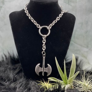 May include: A silver chain necklace with a large silver axe pendant. The necklace is displayed on a black velvet mannequin bust.