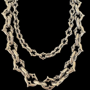 Puede incluir: Un collar plateado con dos hilos de eslabones de cadena ornamentados. Las cadenas tienen un patrón repetitivo de eslabones en forma de diamante con cortes decorativos. El collar se muestra sobre un fondo negro, resaltando el intrincado diseño.