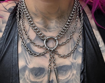 May include: A multi-layered silver-toned metal chain necklace featuring a central circular ring connector. Two shorter chains hang from the ring, each ending in a pointed metal spike pendant. The necklace consists of several interconnected curb chains.