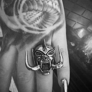 Puede incluir: Un anillo de plata con un diseño detallado de una cabeza de animal gruñendo con cuernos. El anillo se lleva en un dedo con uñas largas pintadas.