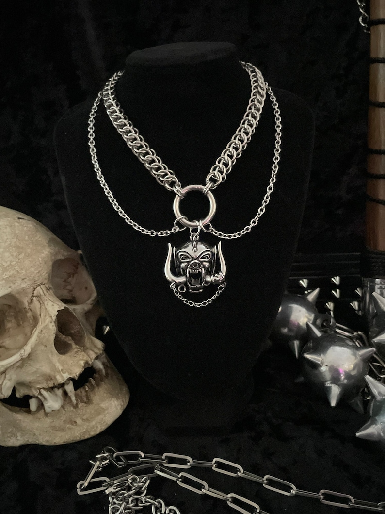 May include: A silver chain necklace with a large silver pendant featuring a snarling animal head with horns. The pendant is attached to a large silver ring.