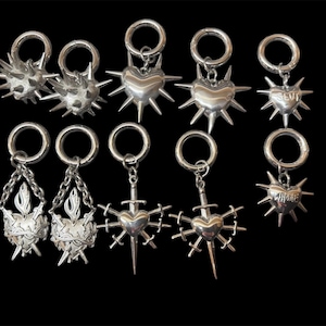 May include: A collection of silver-toned earrings with various designs, including spiked hearts, hearts with swords, and a heart with the word "SLUT". Some earrings feature a chain or a ring attachment.