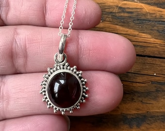 Op de afbeelding: Een zilveren ketting met een donkerrode edelsteen hanger omringd door een zilveren zonnestralen ontwerp.