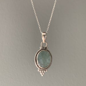 Op de afbeelding: Een zilveren ketting met een ronde, lichtblauwe edelsteen hanger. De hanger is gezet in een zilveren frame met een decoratieve rand.