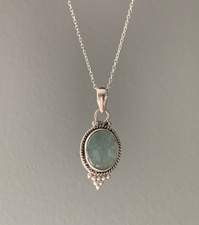 Op de afbeelding: Een zilveren ketting met een ronde, lichtblauwe edelsteen hanger. De hanger is gezet in een zilveren frame met een decoratieve rand.