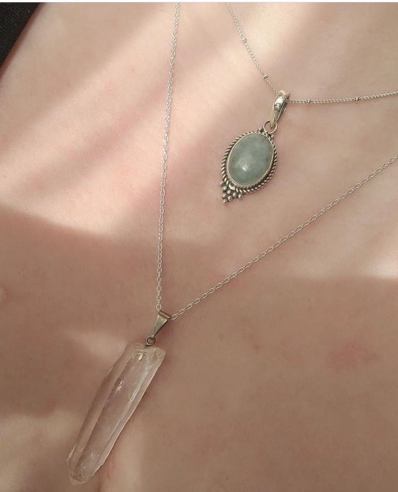 Op de afbeelding: Twee zilveren kettingen met kristallen hangers. De ene hanger is een heldere kwartskristal, de andere is een lichtblauwe aquamarijnkristal.