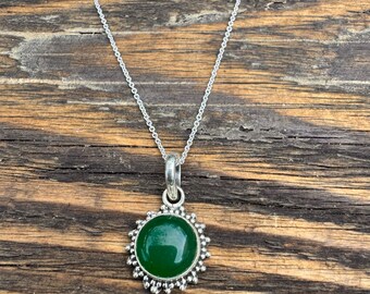 May include: A silver necklace with a round, deep green pendant. The pendant is surrounded by a decorative silver border. The necklace chain is delicate and hangs on a wooden surface.