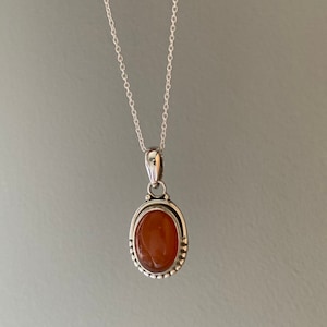 May include: A silver chain necklace with a pendant featuring a carnelian gemstone set in a silver oval bezel with a beaded border.
