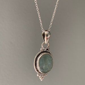Op de afbeelding: Een zilveren ketting met een hanger met een lichtblauwe ovale edelsteen in een zilveren zetting met decoratieve details.