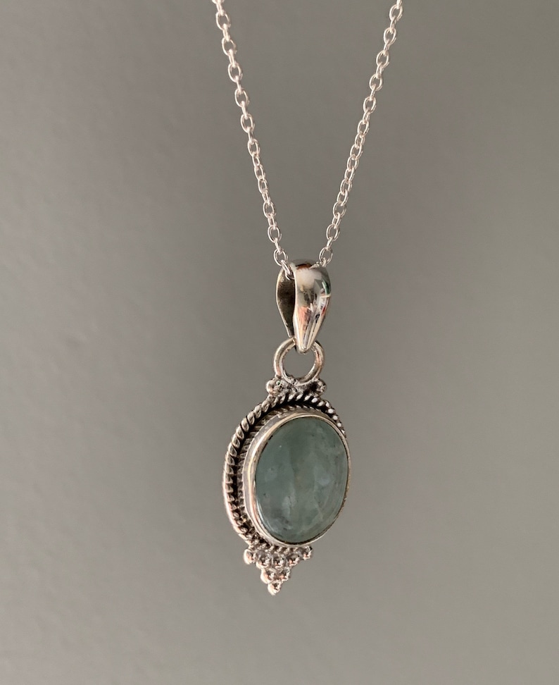 Op de afbeelding: Een zilveren ketting met een hanger met een lichtblauwe ovale edelsteen in een zilveren zetting met decoratieve details.