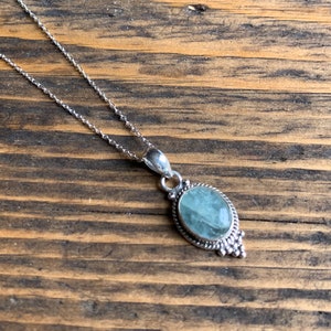 Op de afbeelding: Een zilveren ketting met een hanger met een lichtblauwe ovale edelsteen in een zilveren frame met decoratieve details.