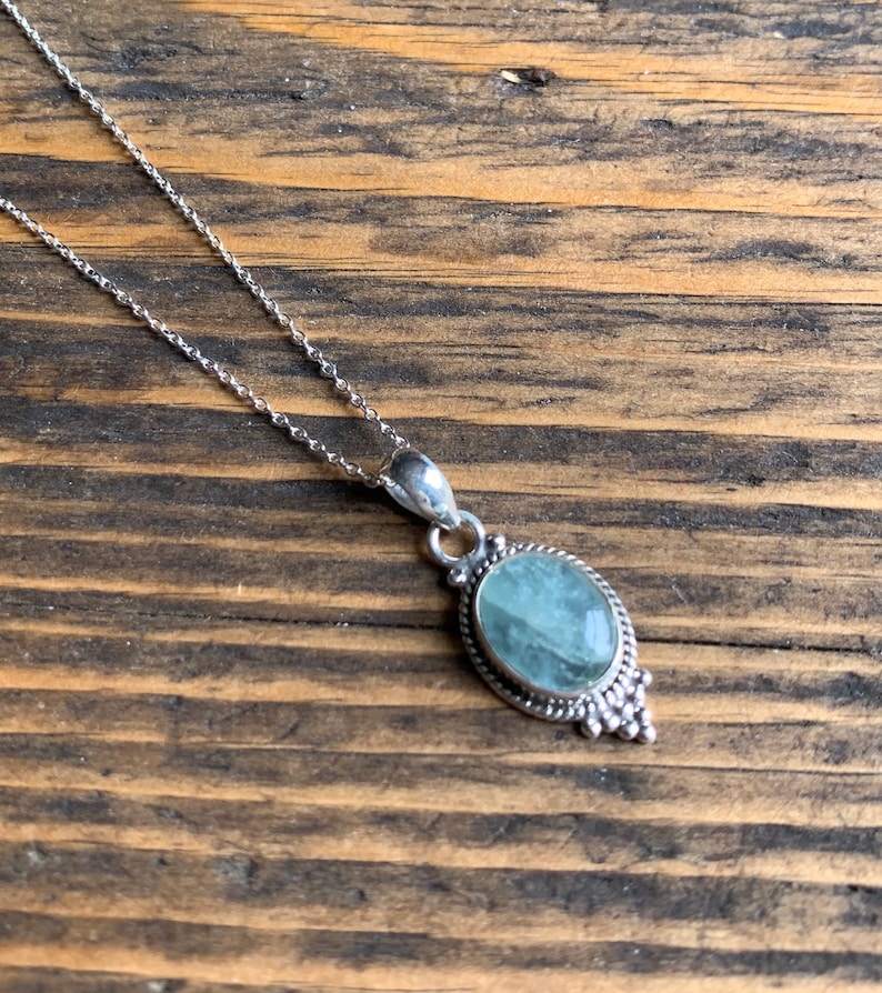 Op de afbeelding: Een zilveren ketting met een hanger met een lichtblauwe ovale edelsteen in een zilveren frame met decoratieve details.