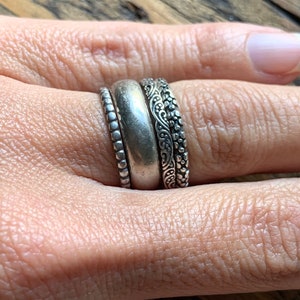 May include: Three silver rings stacked on a finger. The top ring has a delicate floral design, the middle ring is a plain band, and the bottom ring has a beaded design.