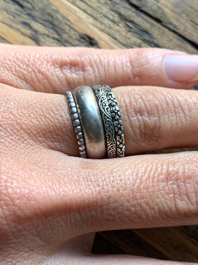 May include: Three silver rings stacked on a finger. The top ring has a delicate floral design, the middle ring is a plain band, and the bottom ring has a beaded design.