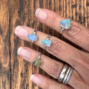 May include: Four silver rings with different coloured opal stones. The rings are on a person's hand.