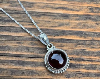 May include: A silver chain necklace with a round pendant featuring a dark red gemstone set in a silver bezel. The pendant is surrounded by a row of small silver beads.