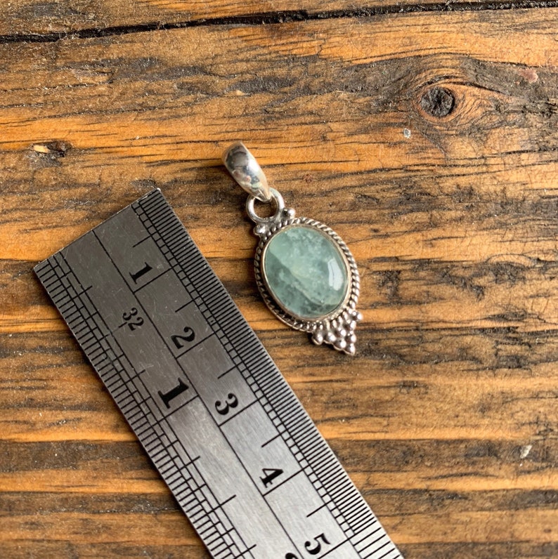 Op de afbeelding: Een zilveren hanger met een lichtblauwe ovale edelsteen gezet in een zilveren bezel. De hanger is ongeveer 2,5 cm lang en wordt getoond naast een liniaal.