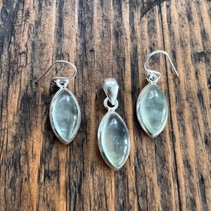 Puede incluir: Un conjunto de pendientes y un colgante de plata con piedras ovaladas de color verde claro.