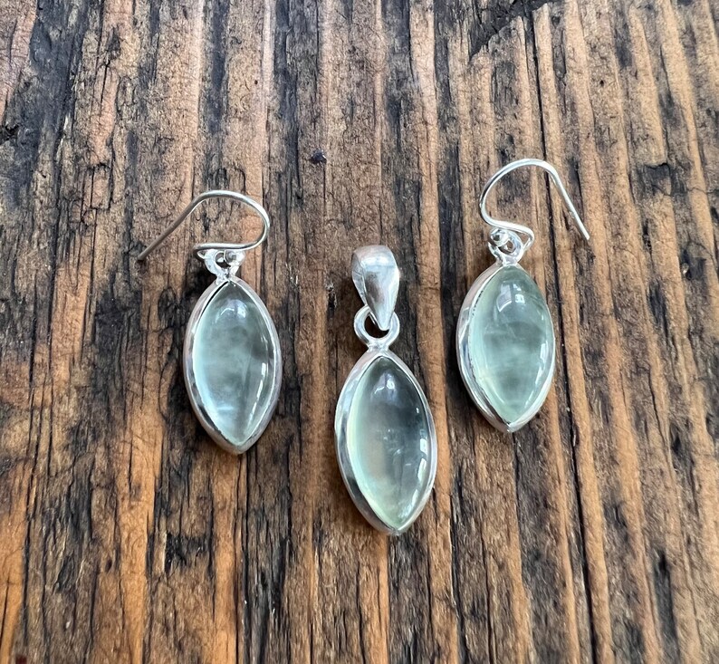 Puede incluir: Un conjunto de pendientes y un colgante de plata con piedras ovaladas de color verde claro.