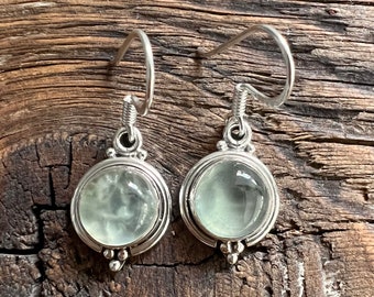 May include: A pair of silver earrings with a round, light green gemstone in each. The earrings have a simple, elegant design.